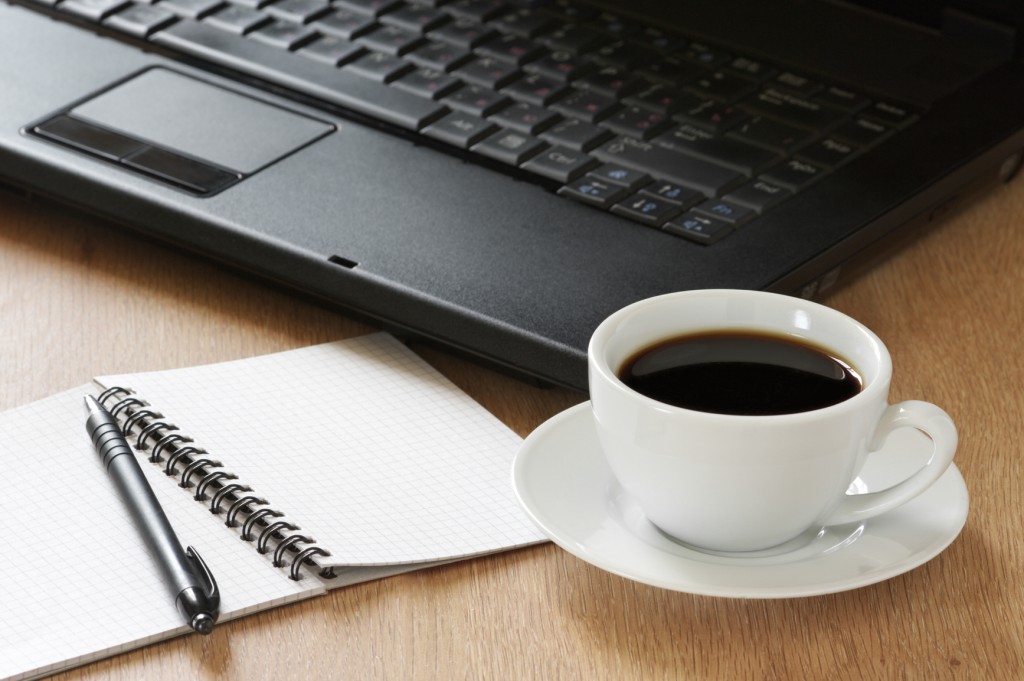 Laptop, notepad, pen and cup of coffee on wooden desk.